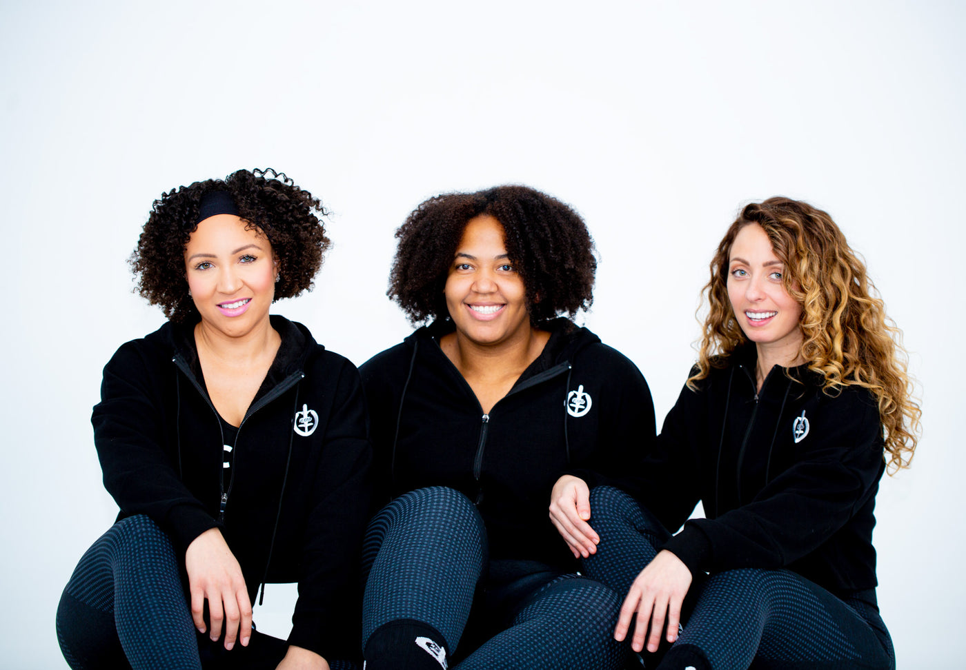 Three diverse women sitting together in ICONI Core Cropped Hoodies in the color black and ICONI Core Leggings in the color Ash.