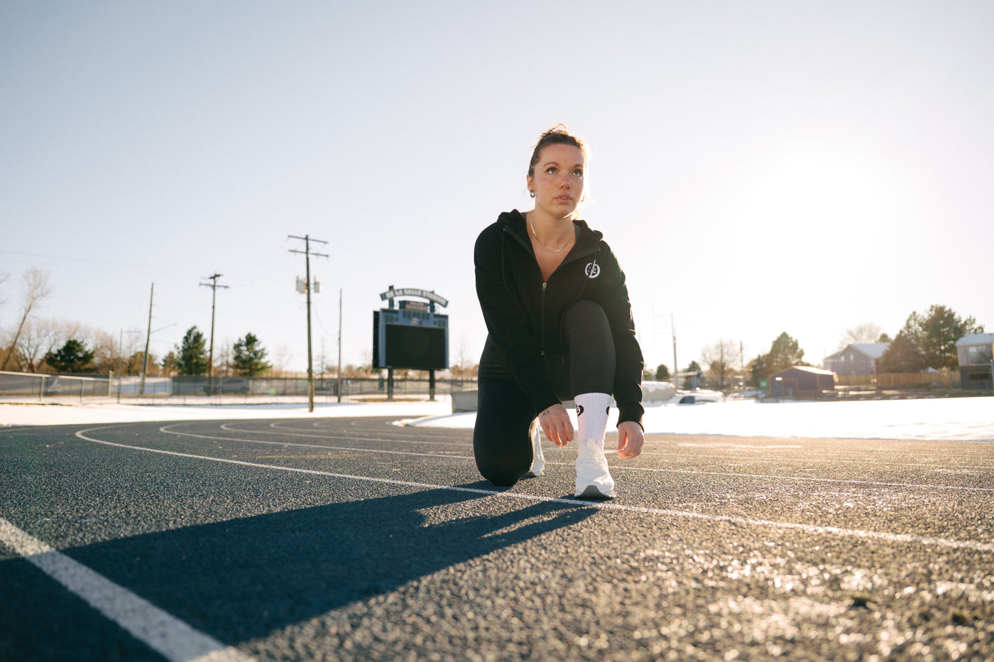 Woman in black hoodie, black leggings, and white socks tying her shoes on a track.