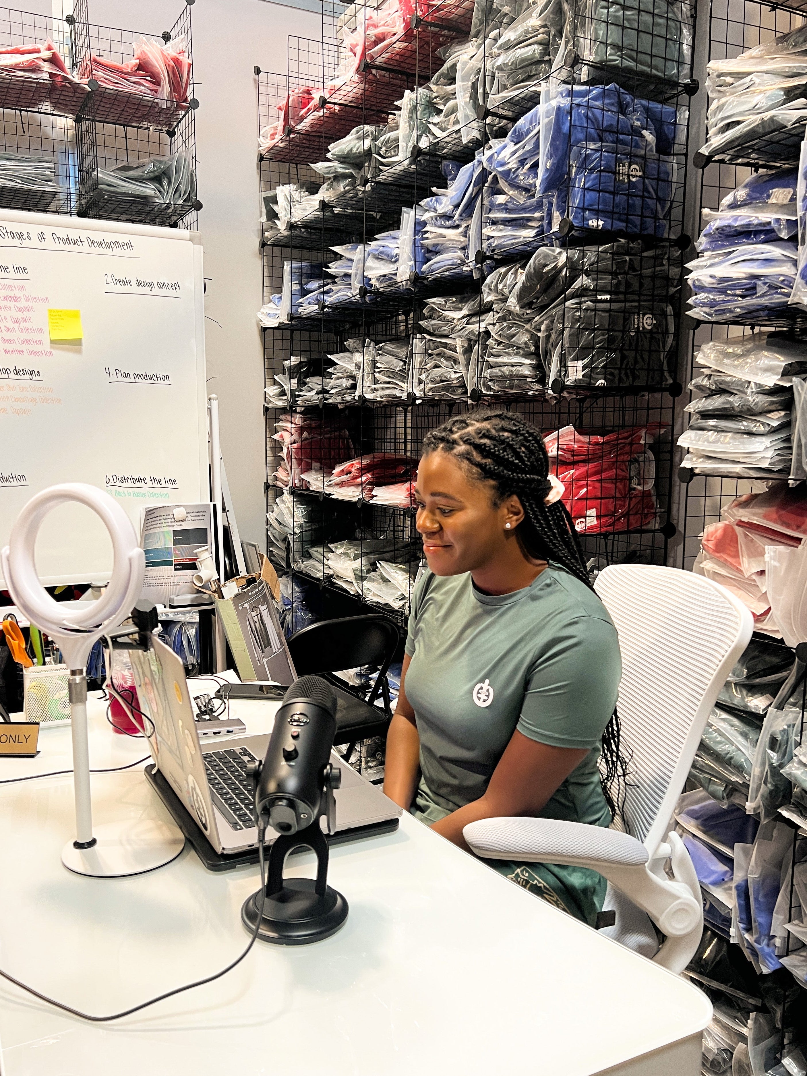 Woman in warehouse sitting at desk on computer with ring light and microphone. Behind her is a wall unit full of leggings.