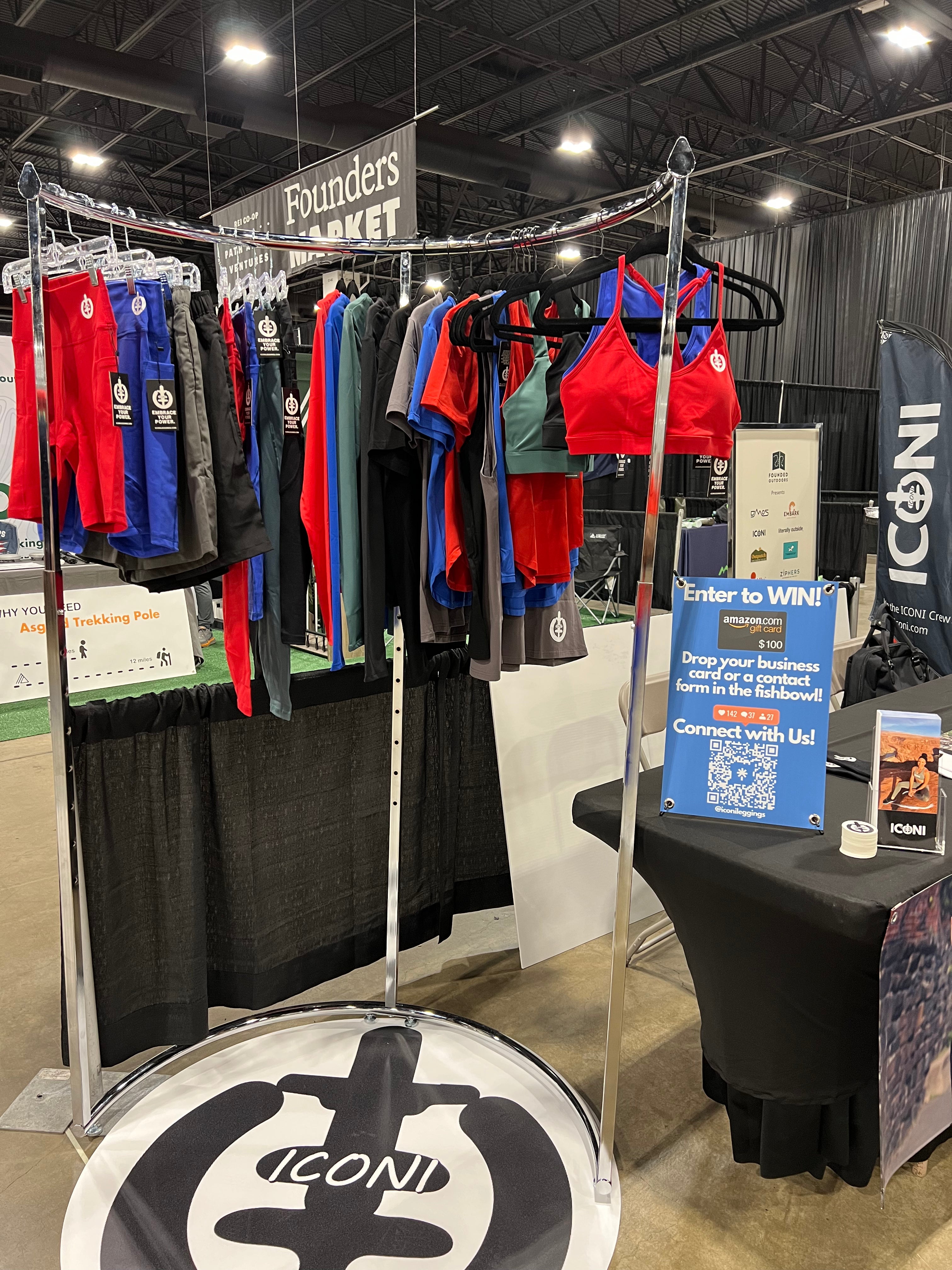 Display of red, jade, blue, and black sports bras, leggings, shorts, and shirts on a silver metal rack with an ICONI floor cover underneath